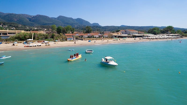 Aerial view of a Mediterranean resort beach with turquoise water, families enjoying a banana boat and white parasols along the shoreline
