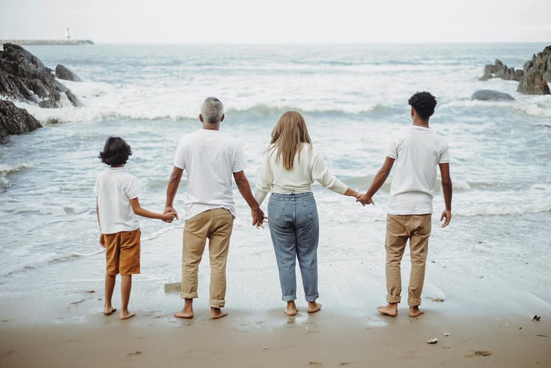 A family of four holds hands facing the ocean on a sunny beach holiday
