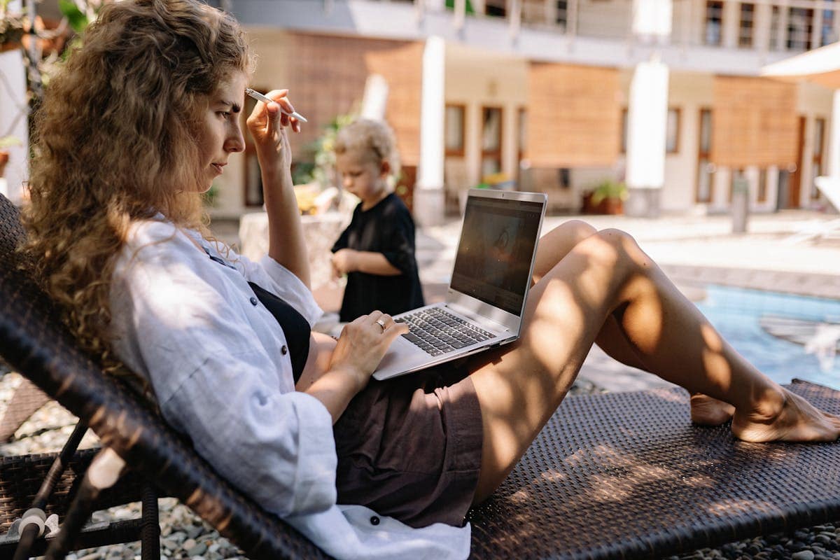 Booking a school holiday deal online Woman relaxing on a sunlounger by a pool with a laptop, planning a holiday deal